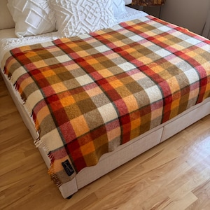 May include: A woven throw blanket featuring a plaid pattern in shades of orange, red, brown, and cream. The blanket has a fringed edge and a small rectangular black label with white text attached to the corner, draped over a bed.
