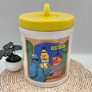 Peut inclure: Un pot à biscuits en céramique blanche avec un couvercle jaune vif muni d'une poignée. L'avant présente une illustration colorée de Cookie Monster, Bert et Ernie de Sesame Street, avec le texte Sesame Street imprimé dans un rectangle jaune.
