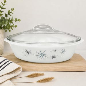 Puede incluir: Una fuente de horno ovalada de vidrio blanco con una tapa de vidrio transparente. El exterior de la fuente presenta un patrón repetitivo de estrellas azul oscuro y pequeños puntos. La fuente tiene asas integradas y un acabado liso y brillante.