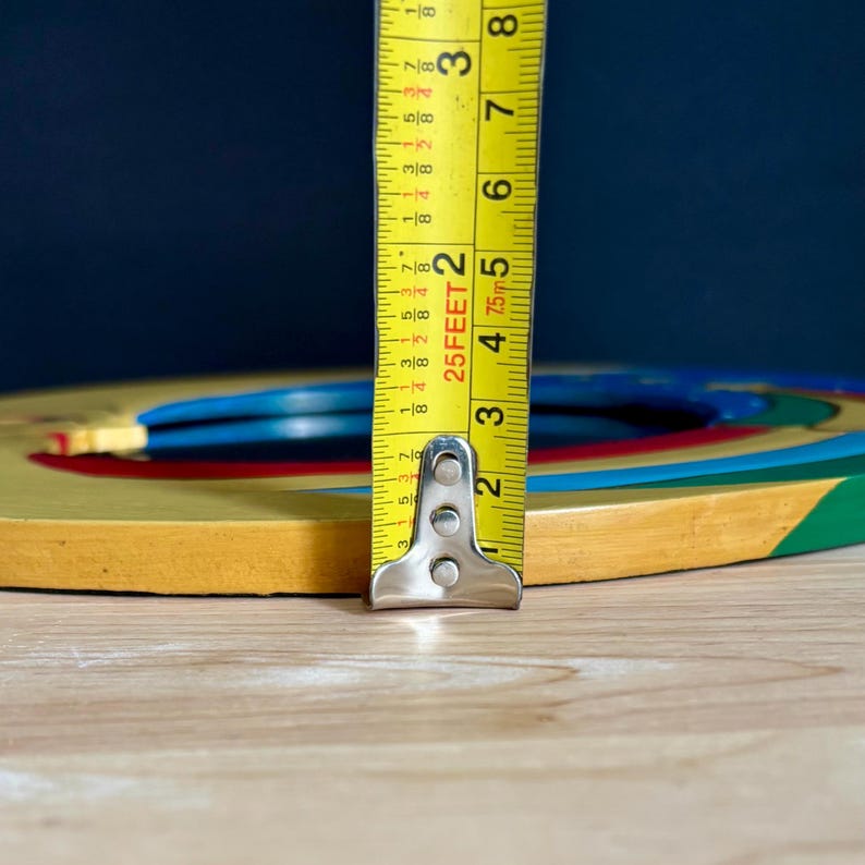 May include: A yellow tape measure with black markings is positioned vertically, showing measurements in inches. The tape measure is placed next to a colorful wooden toy with blue, red, and green accents. The toy appears to be a circular shape.