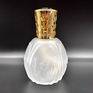 May include: A frosted glass fragrance lamp with a gold-colored, leaf-patterned cap. The round, white glass body features a floral design. The lamp is designed to diffuse fragrance and add a decorative touch to any room.