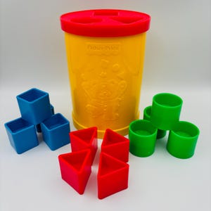 May include: A yellow Fisher-Price plastic shape-sorting canister with a red lid featuring cutouts. The set includes blue square blocks, red triangular blocks, and green cylindrical blocks arranged around the central yellow container.