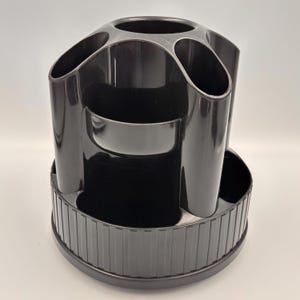 May include: A black desk organizer with multiple compartments for pens, pencils, and other office supplies. The organizer has a round base with a ribbed design and several cylindrical and angled compartments.