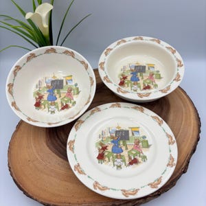 May include: A three-piece ceramic dinnerware set comprising a bowl, a smaller bowl, and a plate, all decorated with a repeating border of brown rabbits and a central illustration of rabbits in a classroom setting with a chalkboard and desks.