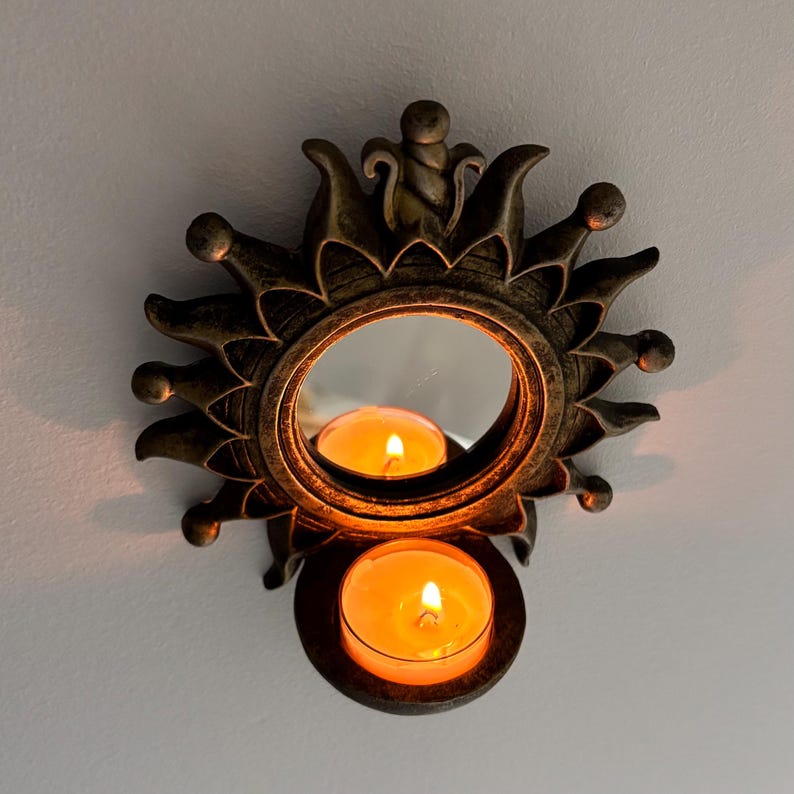 May include: A sun-shaped wall sconce with a dark bronze finish and a central circular mirror. The frame features radiating points and a small, integrated circular holder at the bottom containing a lit orange tea light candle.