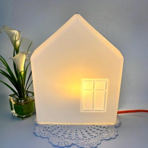 Könnte beinhalten: Eine hausförmige Lampe aus mattiertem weißem Glas, die ein warmes gelbes Licht ausstrahlt. Die Lampe hat ein schlichtes, geprägtes Fensterdesign auf der Vorderseite. Sie steht auf einer weißen Häkeldeckchen neben einer Glasvase mit weißen Calla-Lilien.