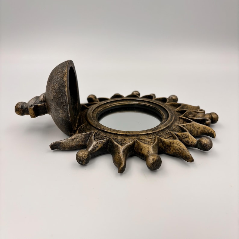 May include: A circular sun-shaped mirror with a distressed bronze-colored finish. The frame features radiating rays with rounded tips. A hinged, semi-spherical compartment is attached to the left side, shown in an open position.