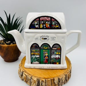 May include: A white ceramic teapot shaped like a building with a cafe storefront. The front features the text Polly's Home Made Teapot Cafe Sweets and Cakes and a cafe teapot. The design includes colourful illustrations of people and baked goods.