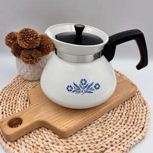May include: A white teapot with a black lid and handle, featuring a blue floral pattern on the front. The teapot has a silver-toned metal band around the neck and a rounded body shape. The design includes a central flower with smaller buds.