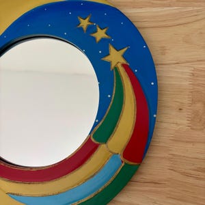May include: A round mirror with a blue frame featuring a shooting star design. The star's trail is composed of red, yellow, green, and blue stripes. Three gold stars are painted on the blue frame, with white dots representing stars.