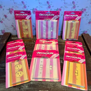 May include: A collection of vintage decorplate clear plastic switch plates and plug plates. The plates feature vertical stripes in shades of pink, yellow, and orange. The packaging is pink with the brand name "decorplate".