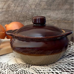 May include: A covered brown ceramic casserole dish with a light tan base and a dark brown lid. The dish has two handles and a small knob on top. The dish is sitting on a white lace tablecloth.
