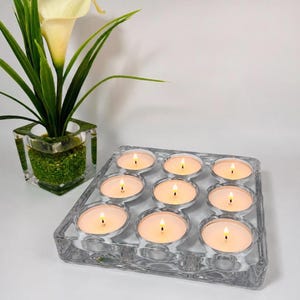 May include: A square clear glass candle holder with nine circular indentations, each containing a lit white tea light candle. Beside it sits a square glass vase filled with green decorative pebbles and a single cream-colored calla lily flower.