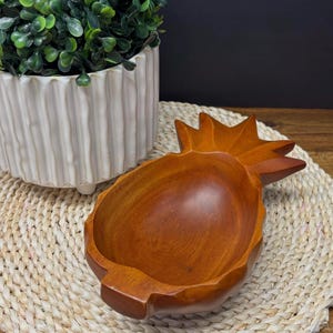 Puede incluir: Un cuenco de madera con forma de piña y un rico acabado marrón. El cuenco se encuentra sobre una esterilla tejida, con una maceta de cerámica blanca y vegetación en el fondo. Ideal para servir aperitivos o como pieza decorativa.
