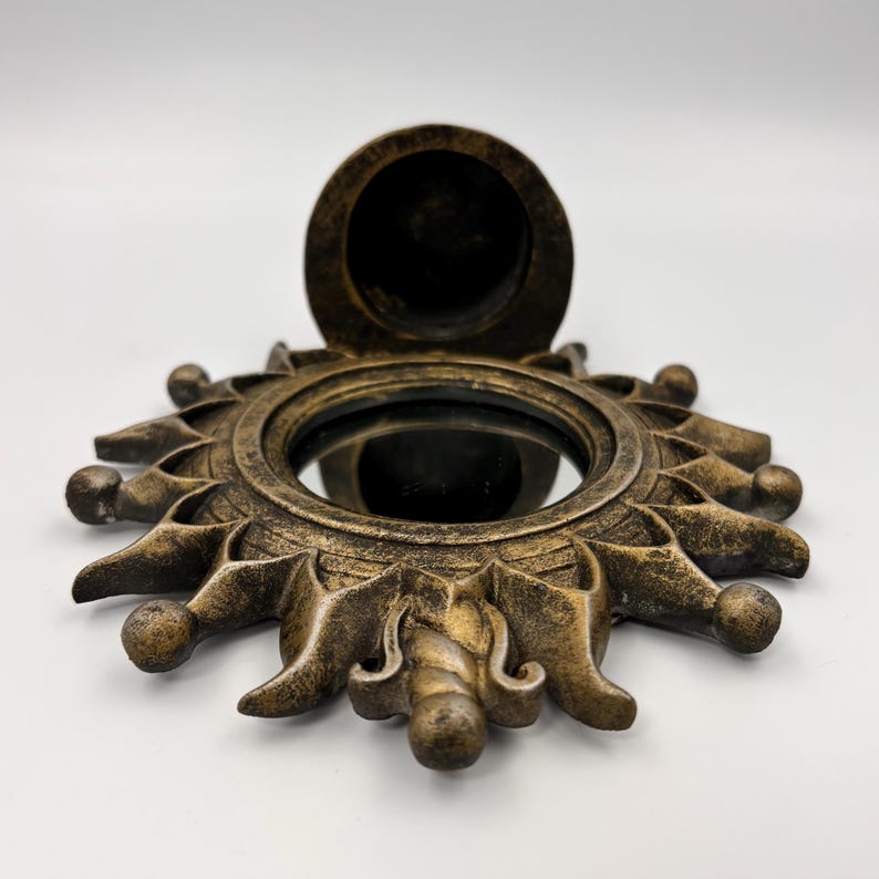May include: A circular wall mirror with a sunburst frame finished in a distressed antique gold color. The frame features radiating pointed rays with rounded tips surrounding a central reflective glass surface and a small circular compartment.