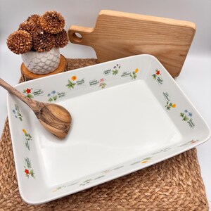 May include: A rectangular white ceramic baking dish featuring a botanical pattern of small colourful wildflowers and Latin text labels along the inner rim. A wooden spoon with a visible grain rests inside the dish, which is placed on a woven mat.