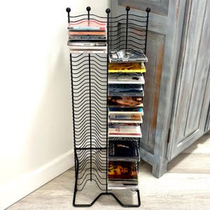 May include: Black metal CD rack with a wavy design. The rack holds multiple CDs, displayed on several shelves. The rack has a decorative top with small ball accents and a sturdy base. The rack is placed next to a light blue cabinet.