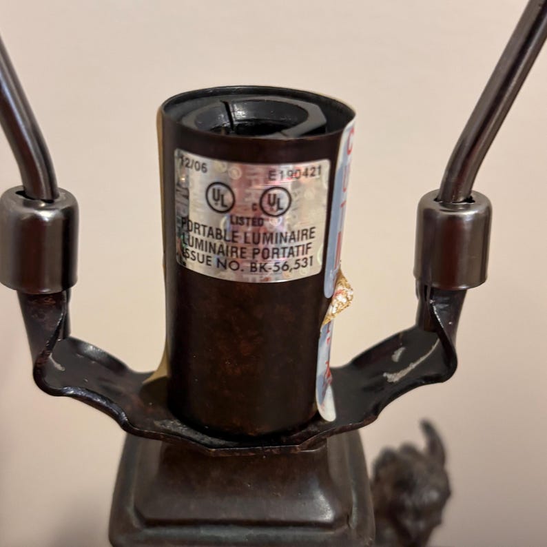 K&ouml;nnte beinhalten: Eine dunkelbraune Lampenfassungsbaugruppe aus Metall mit einem silbernen UL-gelisteten Etikett mit dem Text Portable Luminaire, Luminaire Portatif, Issue No. BK-56,531 und E190421. Die Fassung wird von zwei geschwungenen Metallarmen flankiert.