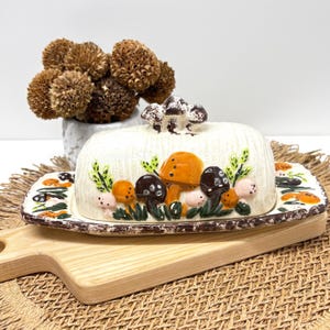 Könnte beinhalten: Eine Keramik-Butterdose mit Deckel, verziert mit bunten Pilzen und grünem Laub. Die Schale ist rechteckig mit einem braunen Rand und steht auf einem Holzschneidebrett. Ein kleiner Strauß Trockenblumen befindet sich im Hintergrund.