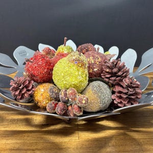 Può includere: Una ciotola decorativa in metallo argentato riempita con frutta e pigne finte assortite. La frutta comprende mele rosse, pere verdi e altri pezzi colorati e strutturati. La composizione è su una superficie di legno.