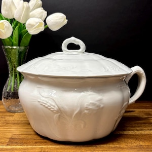 Peut inclure: Un plat de service en céramique blanche avec un couvercle, orné de motifs de blé et de fleurs en relief. Le plat a une poignée sur le côté et une poignée en boucle sur le couvercle. Un vase de tulipes blanches est à gauche.