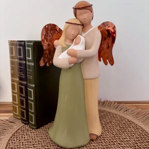 Puede incluir: Una figura de dos adultos con alas de metal color cobre y halos de alambre fino, de pie juntos y sosteniendo a un pequeño bebé envuelto en una tela blanca. Las figuras tienen superficies lisas y mate en tonos verde oliva y beige.