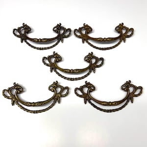May include: Six antique brass drawer pulls with ornate, scrollwork designs. Each pull features a curved handle with a decorative centre and detailed end pieces. The pulls are arranged on a white background.