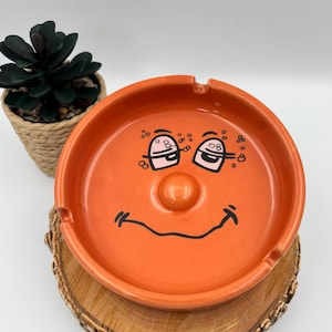 Peut inclure: Un cendrier rond en céramique orange vif orné d'un dessin au trait noir représentant un visage souriant avec deux yeux et une bouche courbée. Le cendrier présente une finition lisse et brillante ainsi que trois encoches sur le bord.