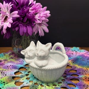 Peut inclure: Un pot de fleurs en céramique blanche en forme de panier, avec deux chats sur le devant. Le pot a une poignée et un motif de panier tressé. Un vase de fleurs violettes est en arrière-plan.