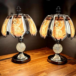 Peut inclure: Deux lampes de table décoratives avec un motif de tigre sur l'abat-jour. Les lampes ont une base sphérique transparente et une monture en métal noir. Les abat-jours sont de couleur orange chaud avec une illustration de tigre. Les lampes sont sur une surface en bois.
