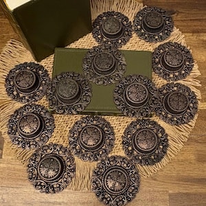 May include: A collection of antique bronze-colored decorative buttons with intricate floral and geometric designs. The buttons are arranged on a woven placemat, with a green book in the background. The buttons have a raised center and detailed edges.