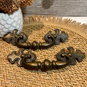 May include: Two antique bronze-colored drawer pulls with ornate, stylized designs. Each pull features a central handle with decorative end pieces. The pulls are displayed on a woven, textured surface, suggesting a vintage or rustic aesthetic.