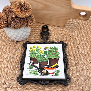 May include: Black cast iron trivet with a ceramic tile featuring potted herbs and a mortar and pestle. The tile has a white background with illustrations of yellow flowers, green herbs, and red berries. The trivet has a decorative handle.