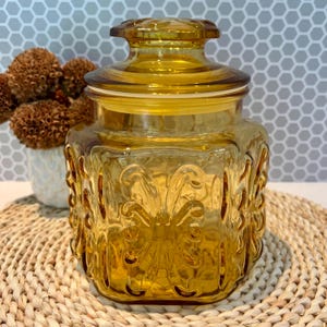 Puede incluir: Un tarro de vidrio ámbar vintage con tapa. El tarro tiene forma cuadrada con diseños florales en relieve. La tapa redonda encaja perfectamente. El tarro está sobre un salvamanteles tejido.