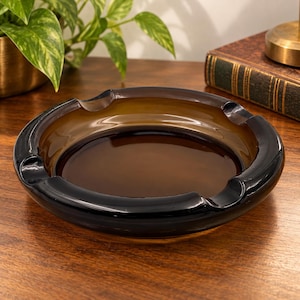 May include: A round glass ashtray featuring a deep amber-colored center basin and a thick, dark black outer rim. The design includes three evenly spaced notches along the perimeter, providing a functional structure for resting items securely.