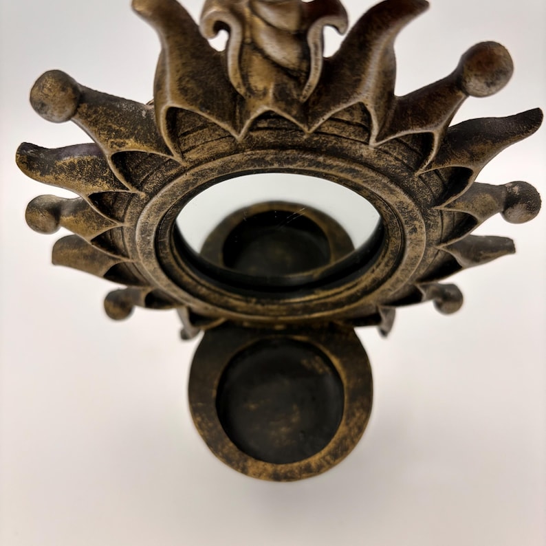 May include: A wall-mounted candle holder featuring a circular mirror framed by a sunburst design with pointed rays. The frame has a distressed bronze finish and includes a small, circular platform at the base for holding a single candle.