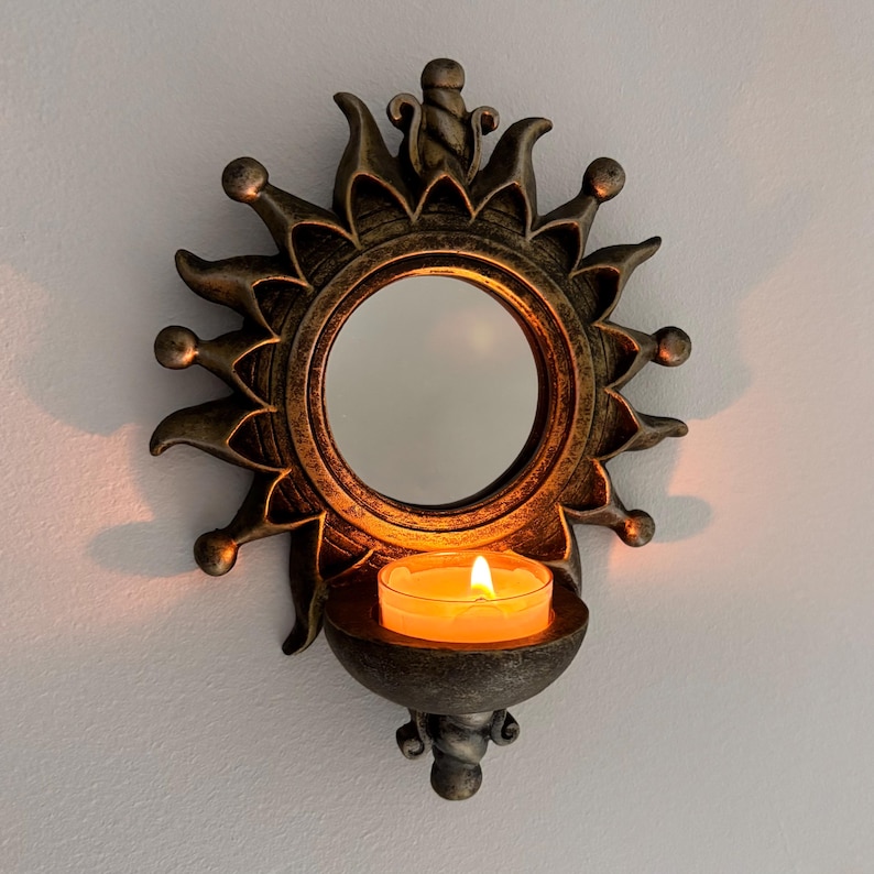 May include: A wall-mounted candle holder featuring a circular mirror framed by a sunburst design in a dark bronze finish. A small glass votive cup sits in the base, holding a lit tea light candle with a warm, glowing orange flame.