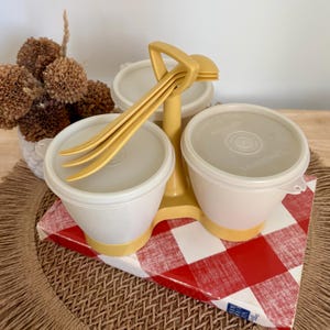 Könnte beinhalten: Vintage Tupperware-Servierset mit drei weißen Schalen, gelben Deckeln und einem gelben Ständer. Das Set enthält gelbes Serviergeschirr und steht auf einer rot-weiß karierten Box.