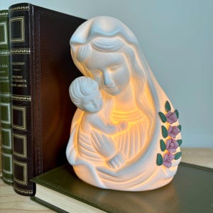 Könnte beinhalten: Eine weiße Keramikfigur der Madonna mit Kind mit sanftem Innenlicht. Die Figur hat einen drapierten Schleier und ist mit rosa Rosen und grünen Blättern verziert. Die Statue steht auf einem grünen Buch.