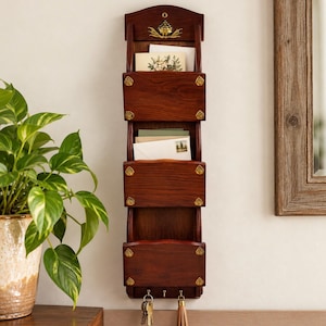 May include: A vertical wall-mounted wooden mail organiser with three storage pockets and three metal hooks at the bottom. The dark brown wood features gold-toned metal accents on the corners and a decorative gold emblem at the top.