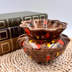 May include: A two-piece ceramic bowl set with a brown glaze and colourful accents of red, yellow, and black. The top bowl has a scalloped edge, and the bottom bowl has a textured surface. The set is placed near a stack of vintage books.