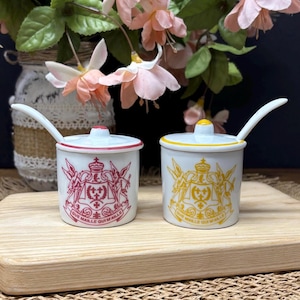 Puede incluir: Dos tarros de condimentos de cerámica blanca con tapas y cucharas. Un tarro tiene un diseño rojo, el otro un diseño amarillo. Ambos tarros tienen un diseño de escudo y el texto "QUE MAILLE QUI MAILLE".