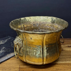 Puede incluir: Una maceta de latón vintage con textura martillada y diseño acanalado. La maceta presenta asas con cabeza de león y un borde decorativo. El latón tiene un tono dorado cálido, ideal para flores o plantas.