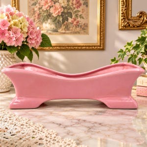 May include: A glossy pink ceramic planter shaped like a miniature clawfoot bathtub with a curved, elongated silhouette and four sturdy feet. The vessel features a smooth, solid-coloured finish and an open top, designed for holding small floral arrangements.