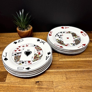 May include: A set of white ceramic plates with a playing card design. Each plate has a black rim and a central image of playing cards, including the Ace of Spades and other suits. The plates are stacked on a wooden surface.