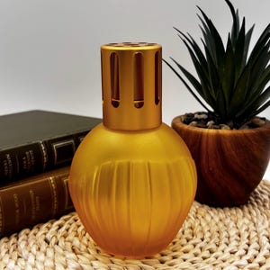 May include: A gold-colored, spherical fragrance lamp with a cylindrical top. The lamp has a ribbed design on the lower portion and sits on a woven mat. A potted plant and books are in the background.