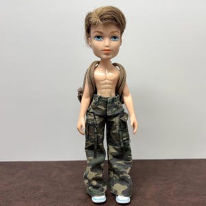 Könnte beinhalten: Eine Bratz-Puppe mit braunen Haaren und blauen Augen, die eine Camouflage-Cargohose und weiße Turnschuhe trägt. Die Puppe hat einen Rucksack und ist ohne Hemd, wobei ein geformter Torso zur Geltung kommt. Die Puppe steht vor einem schlichten Hintergrund.