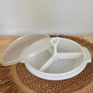 Peut inclure: Un plat de service blanc divisé avec un couvercle transparent. Le plat comporte trois compartiments et une poignée au centre. Le couvercle est partiellement ouvert, révélant l'intérieur. Le plat est sur un tapis tissé.