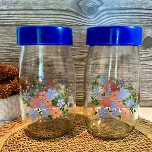 Peut inclure: Deux bocaux en verre transparent avec des couvercles bleus. Les bocaux sont décorés d'un motif floral rose, bleu et vert. Les bocaux sont posés sur un tapis tissé.