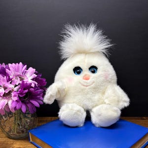 May include: A white plush toy with a fluffy head, blue eyes, and a pink nose and cheeks. The toy is sitting on a blue book. A vase of purple flowers is to the left of the toy.